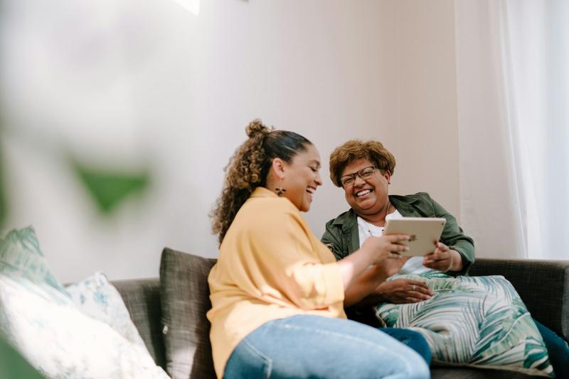 A younger woman and an older woman sit close together on a sofa, laughing as they look at a tablet.