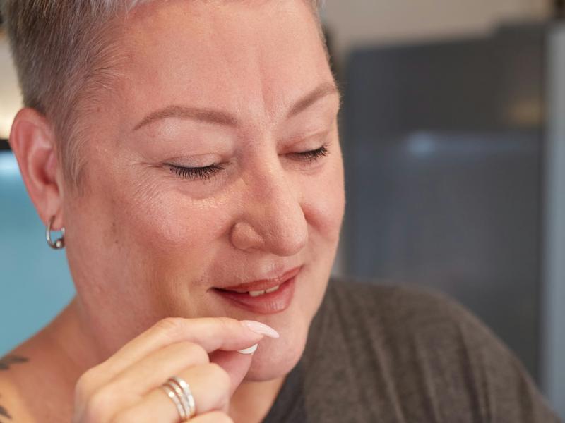 A smiling woman with short grey hair takes a tablet, holding a glass of water in her other hand.