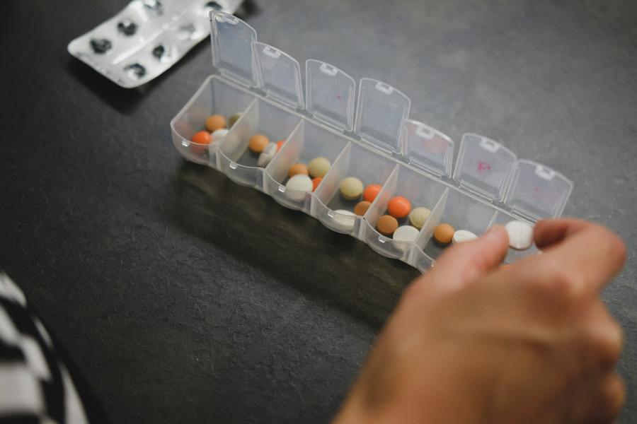 A hand reaches toward an open weekly pill organizer on a dark kitchen table.