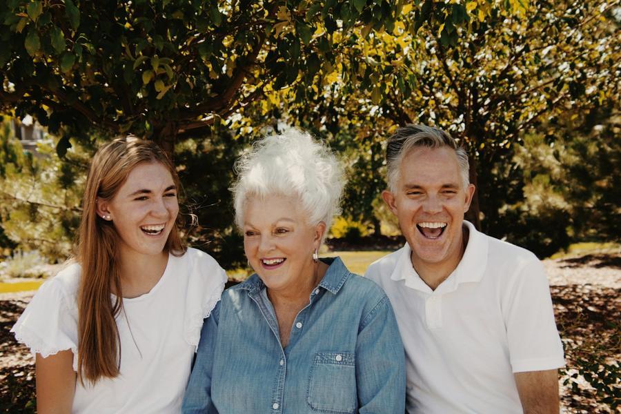 A smiling older woman with white hair sits outdoors between a younger man and a teenage girl, all three laughing.